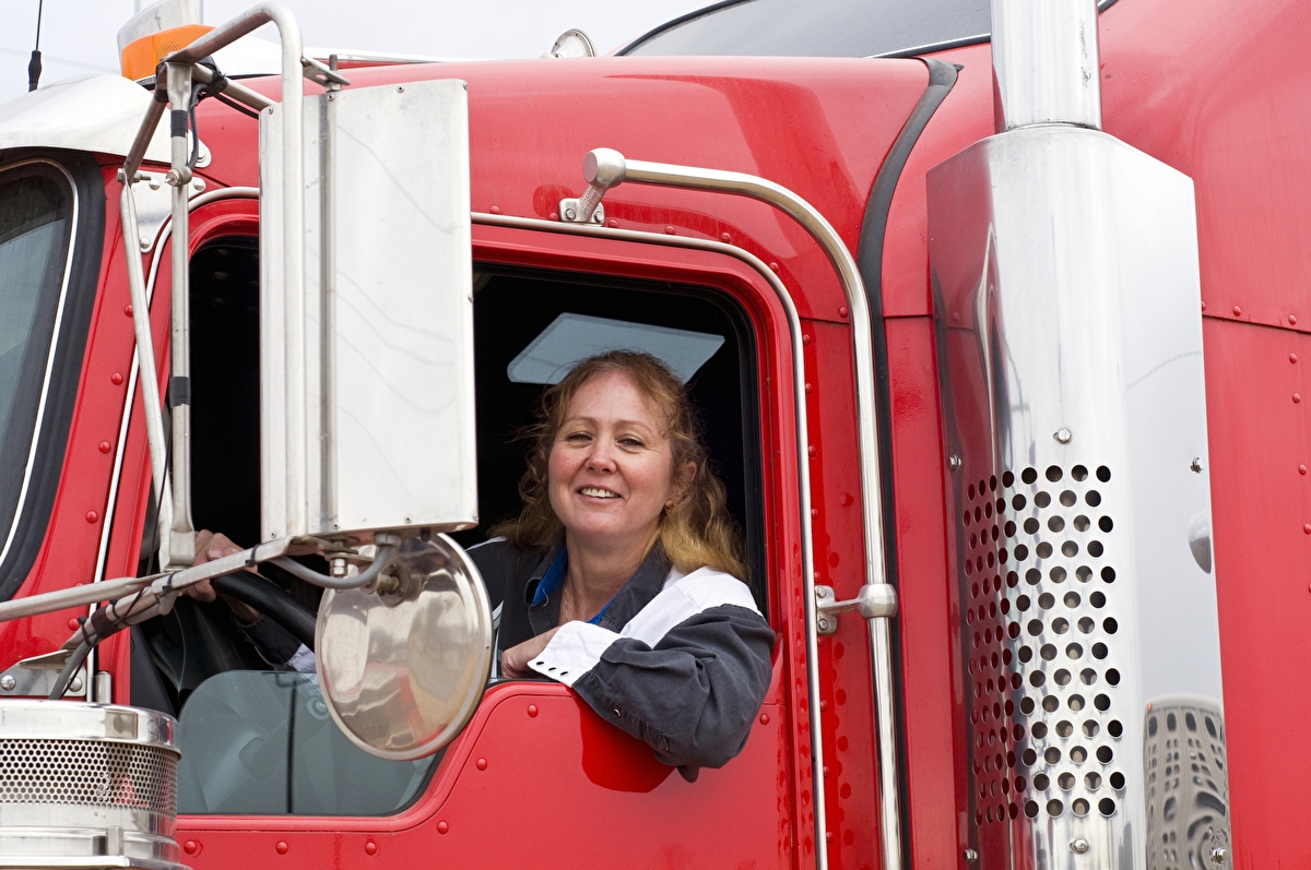 A women sits behind the wheel of a semi truck and looks out its window.
