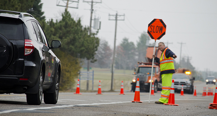 A person in a safety vest holds up a SLOW sign in a roadway work zone as a car approaches.