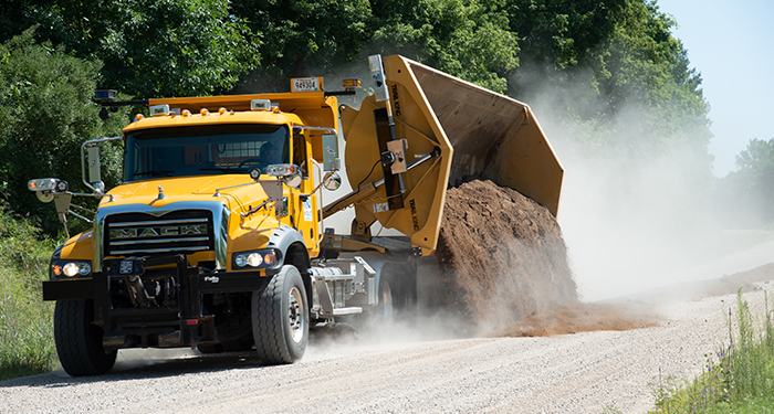 A public works truck dumps dirt onto a gravel road.