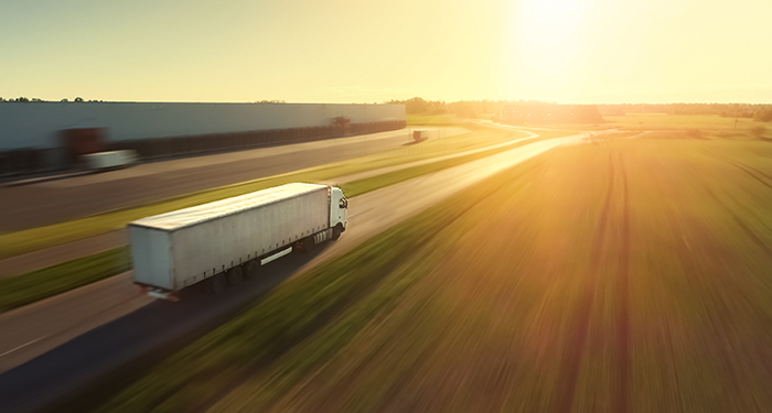 A semi truck drives down a highway into the sun.