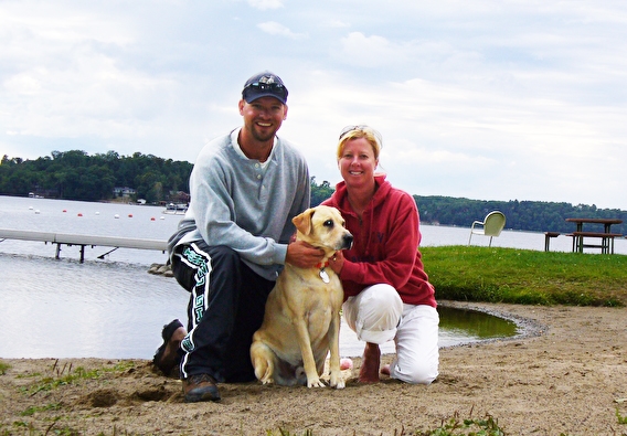 A man and woman kneel on a beach with a yellow lab dog between them.