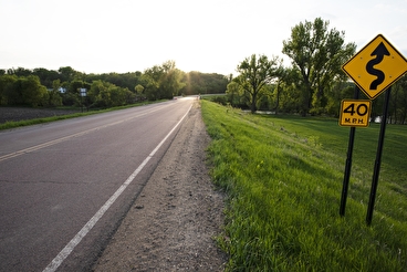 A paved road with a curve-ahead sign in the grass indicating cars should drive no faster than 40 mph.