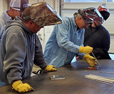 People lean over a worktable wearing welding helmets and gloves.